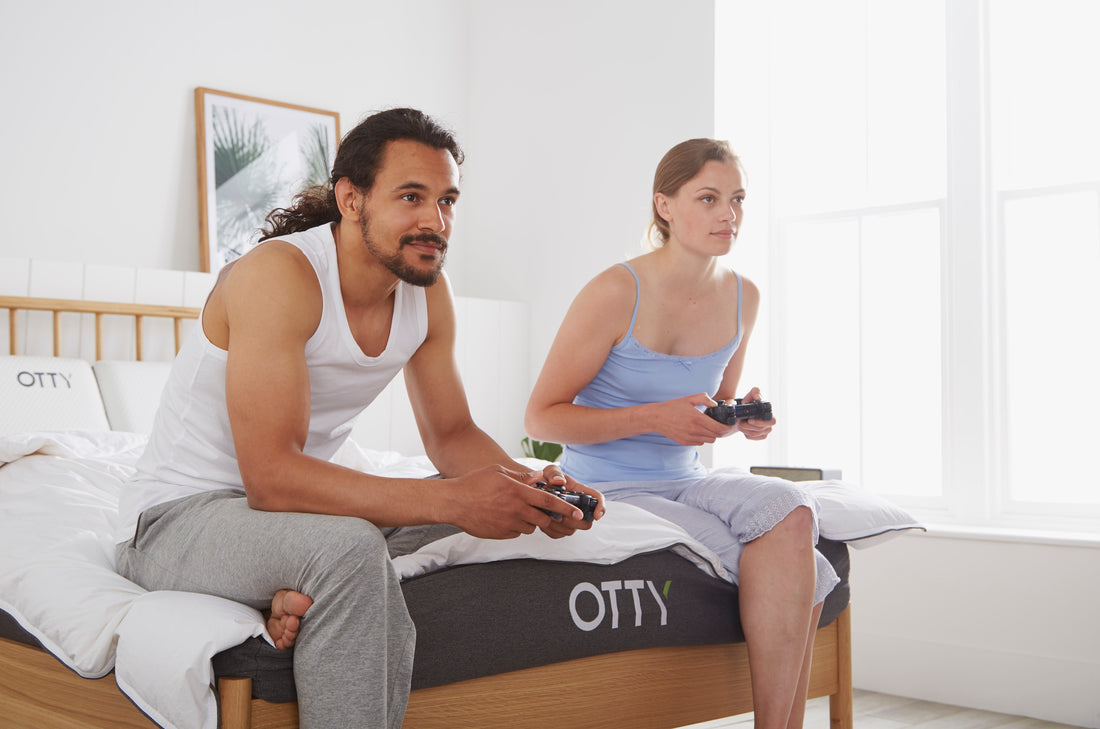 couple sat on top of a hybrid mattress enjoying a game on their playstation. (it's fifa by the way)